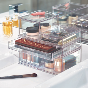 L'organizer modulare HOME EDIT con cassetto di iDesign, disponibile in diverse dimensioni, pieno di palette, fondotinta, rossetti e pennelli, sta sul ripiano bianco del bagno insieme a flaconi di profumo e un pennello da trucco.