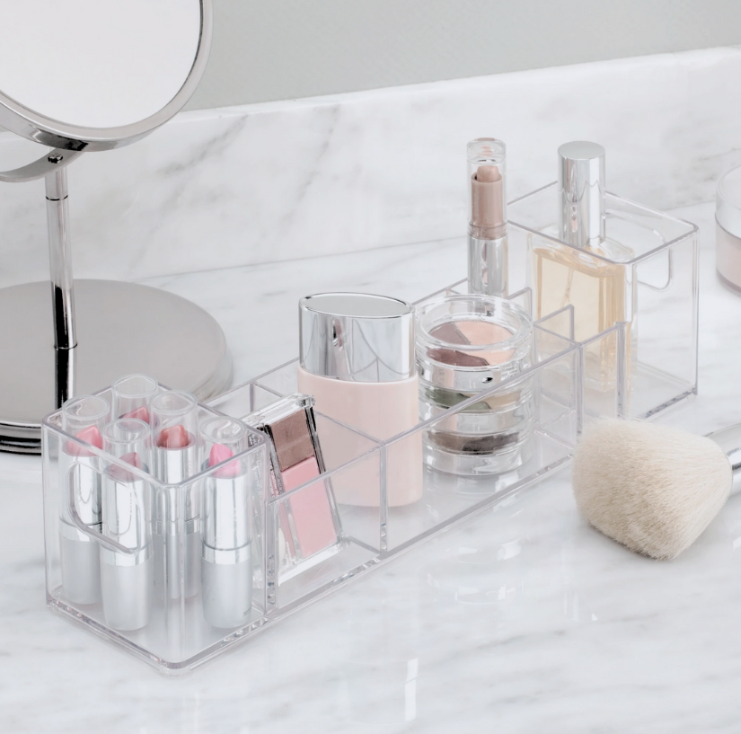 Klarer Kosmetik-Organizer mit Lippenstiften, Puderdosen und Flakons auf weißer Marmoroberfläche neben Spiegel.
