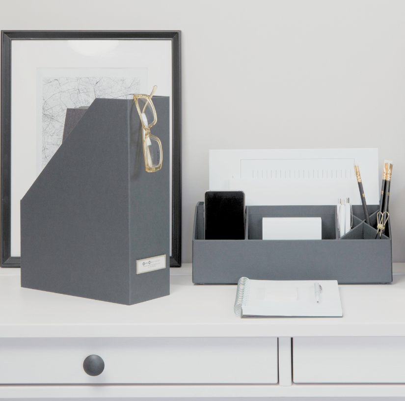 Grauer Stehordner mit Brille steht neben Schreibtisch-Organizer mit Handy und Stiften vor Wandbild.