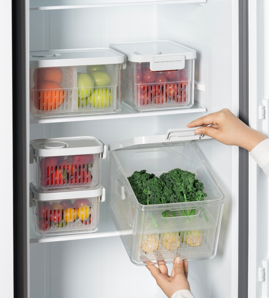 A person stores green leafy vegetables in a transparent DATEMATE KLAR White freshness box with a shelf life timer from BNB x VCN in the refrigerator. Thanks to their stackable design, similar boxes keep fruit and vegetables neatly organized on the shelf.