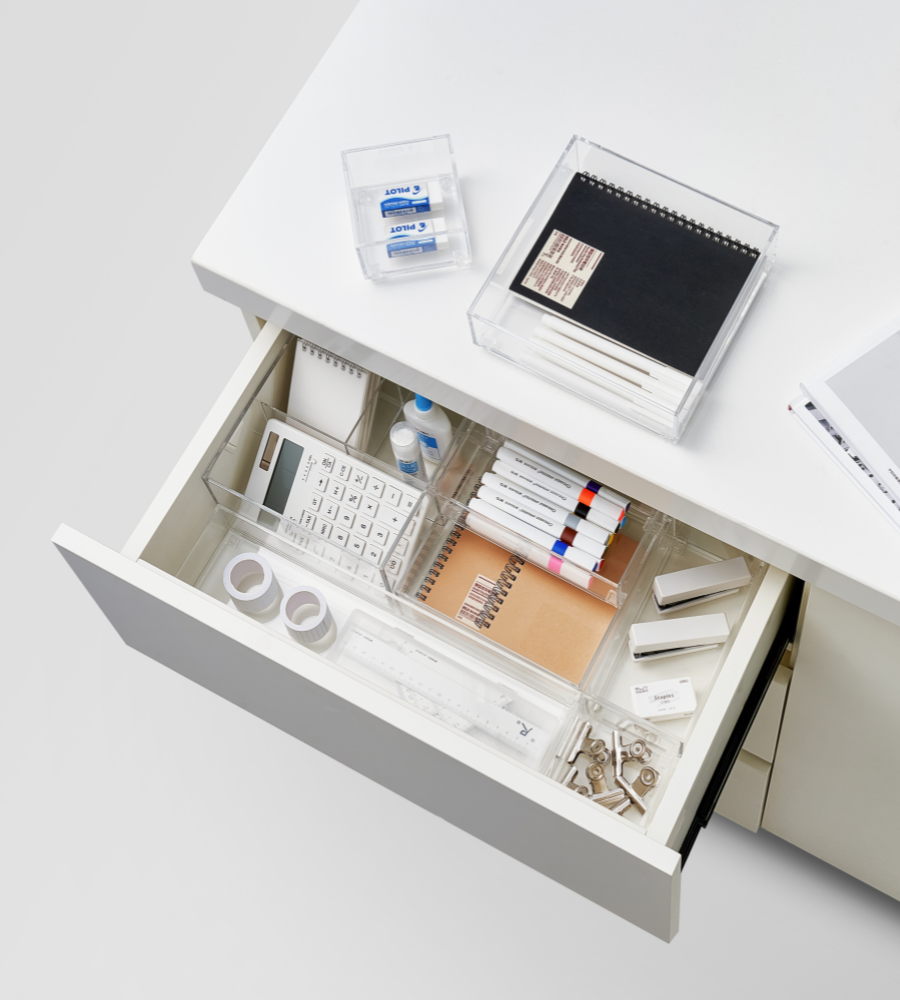 In einer offenen Schreibtischschublade mit dem BNB x VCN Schubladen-Organizer-Set SLIDE (5-teilig) sind Marker, Hefte, Taschenrechner, Korrekturband, Stecknadeln, Klebebandrollen, Lineal und Flaschen übersichtlich untergebracht; Radiergummis und gestapelte Hefte ruhen in durchsichtigen Schreibtischbehältern.