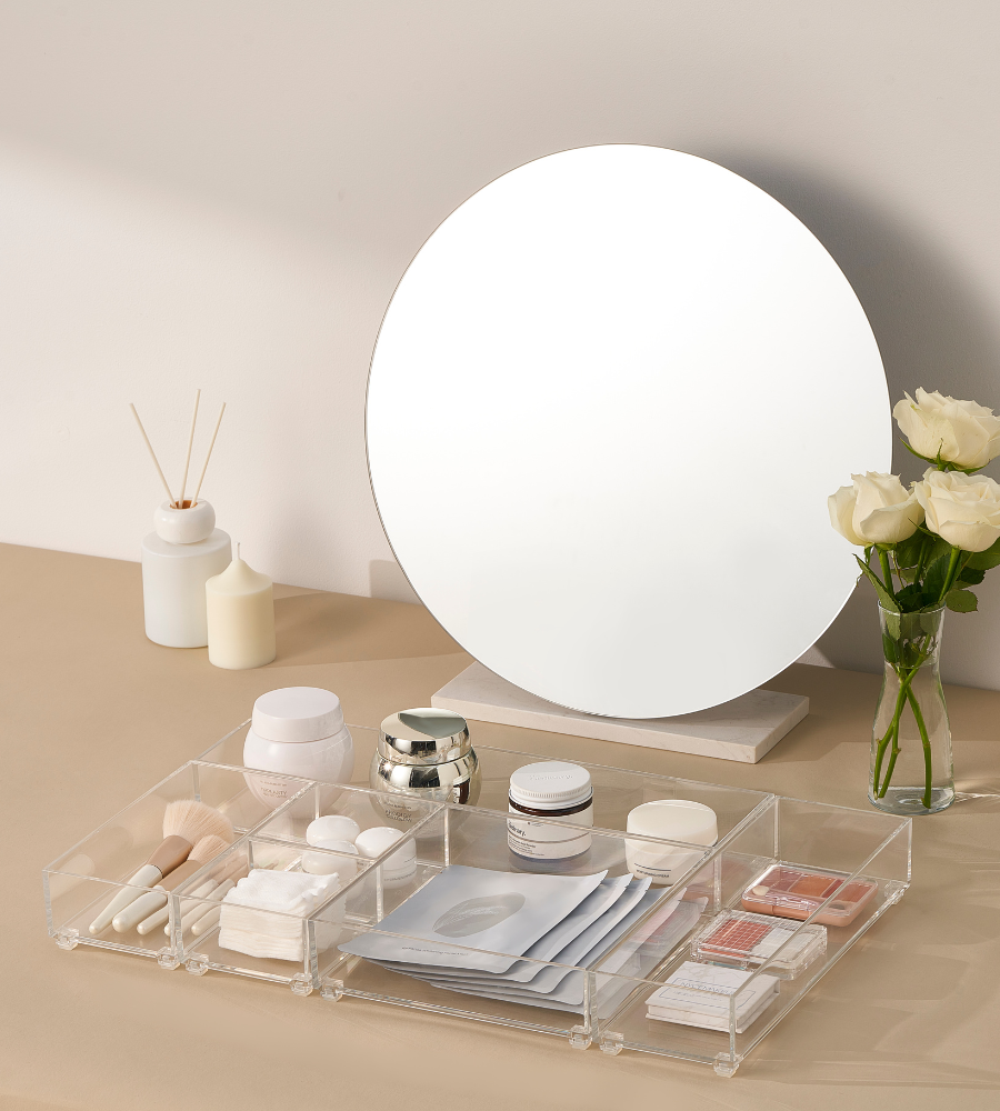 A round mirror stands on a beige washbasin with white roses and candles. Makeup brushes, creams, skincare products, and sheet masks are organized in an organizer and the BNB x VCN drawer organizer set SLIDE (5 pieces).