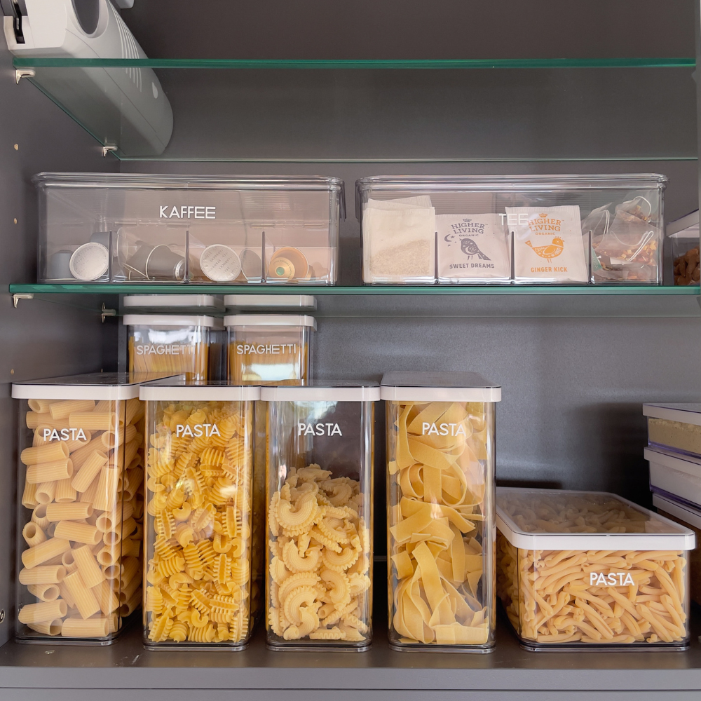 Behälter mit Kaffeekapseln, Teebeuteln und Nudeln im Regal, Texte: „KAFFEE“, „TEE“, „PASTA“, „SPAGHETTI“.