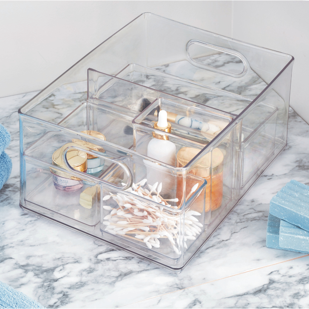 Organizer trasparente con prodotti cosmetici e cotton fioc su un tavolo di marmo, accanto ad asciugamani e sapone.