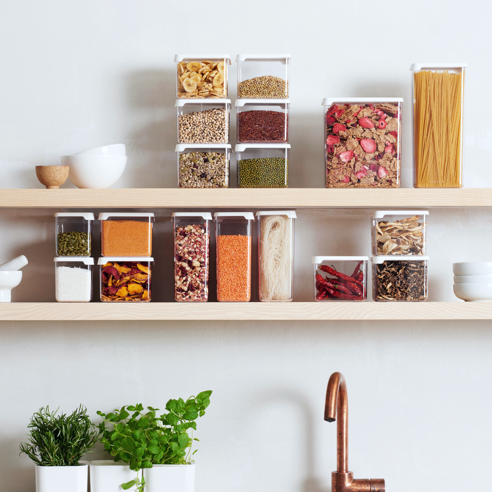 Orthex SmartStore clear storage jars—white in color—filled with grains, pasta, and spices, are neatly arranged on two wooden shelves; herb pots sit on the bottom shelf, and a copper faucet is located below them.