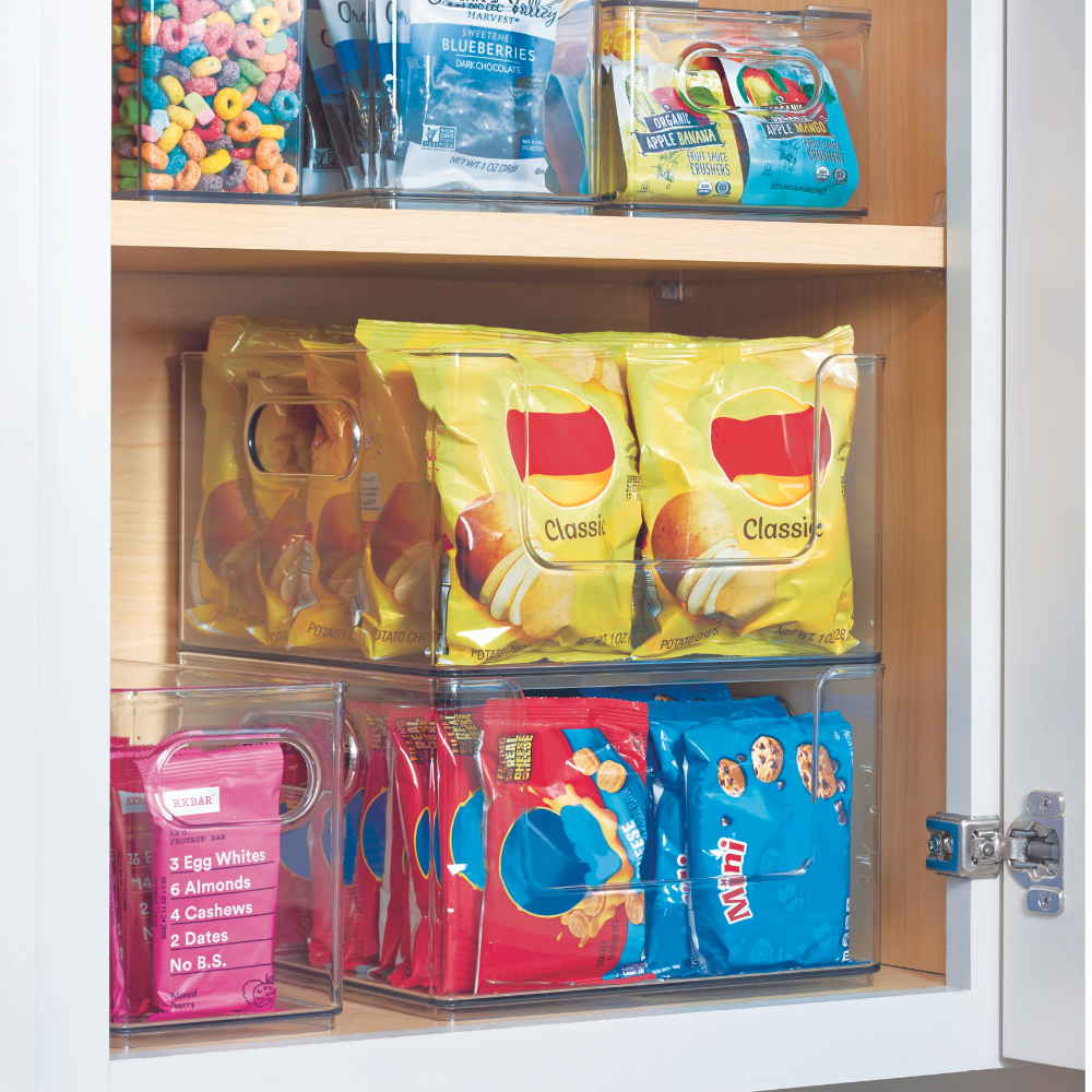 Ein Speisekammerregal mit dem HOME EDIT Aufbewahrungsbehälter KLAR - OpenFront von iDesign hält bunte Snack-Tüten - Chips, Kekse, Müsli und eine Proteinriegel-Box - in drei Organizer-Kunststoffbehältern ordentlich gestapelt im Schrankinneren aufbewahrt.