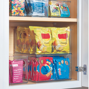 Ein Speisekammerregal mit dem HOME EDIT Aufbewahrungsbehälter KLAR - OpenFront von iDesign hält bunte Snack-Tüten - Chips, Kekse, Müsli und eine Proteinriegel-Box - in drei Organizer-Kunststoffbehältern ordentlich gestapelt im Schrankinneren aufbewahrt.