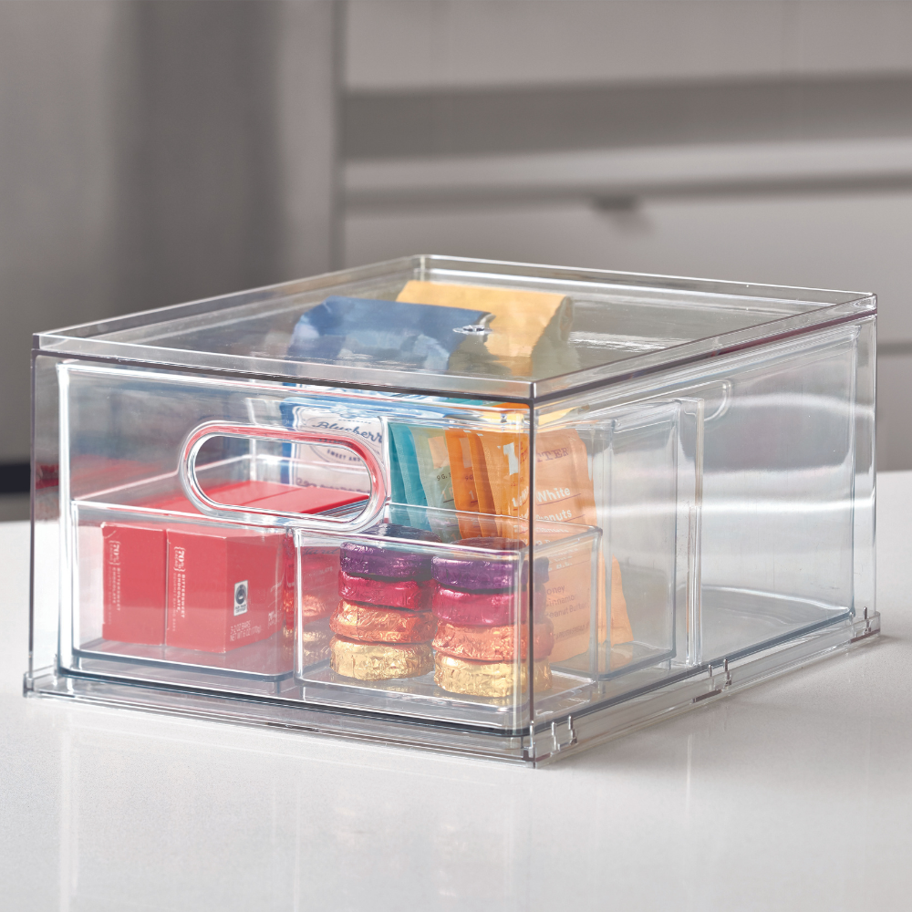 Eine durchsichtige iDesign HOME EDIT - Aufbewahrungsbox Schublade KLAR (35x26,7x19,7cm) mit Henkel, gefüllt mit verschiedenen bunten Snacks, steht auf einer weißen Arbeitsplatte in einer modernen Küche.